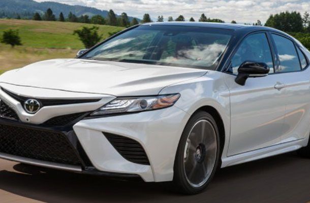 A white Toyota sedan driving on a road, with a scenic landscape of green fields and trees in the background.