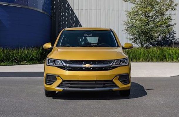 A yellow Chevrolet sedan parked on a road, with a modern building featuring blue and yellow accents and greenery in the background.