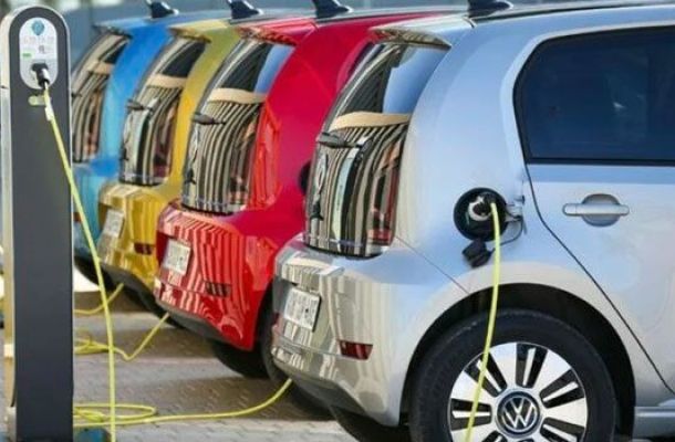 A row of colorful electric cars, including a silver Volkswagen, charging at electric vehicle charging stations, with vibrant hues of blue, yellow, red, and silver creating a visually striking scene.