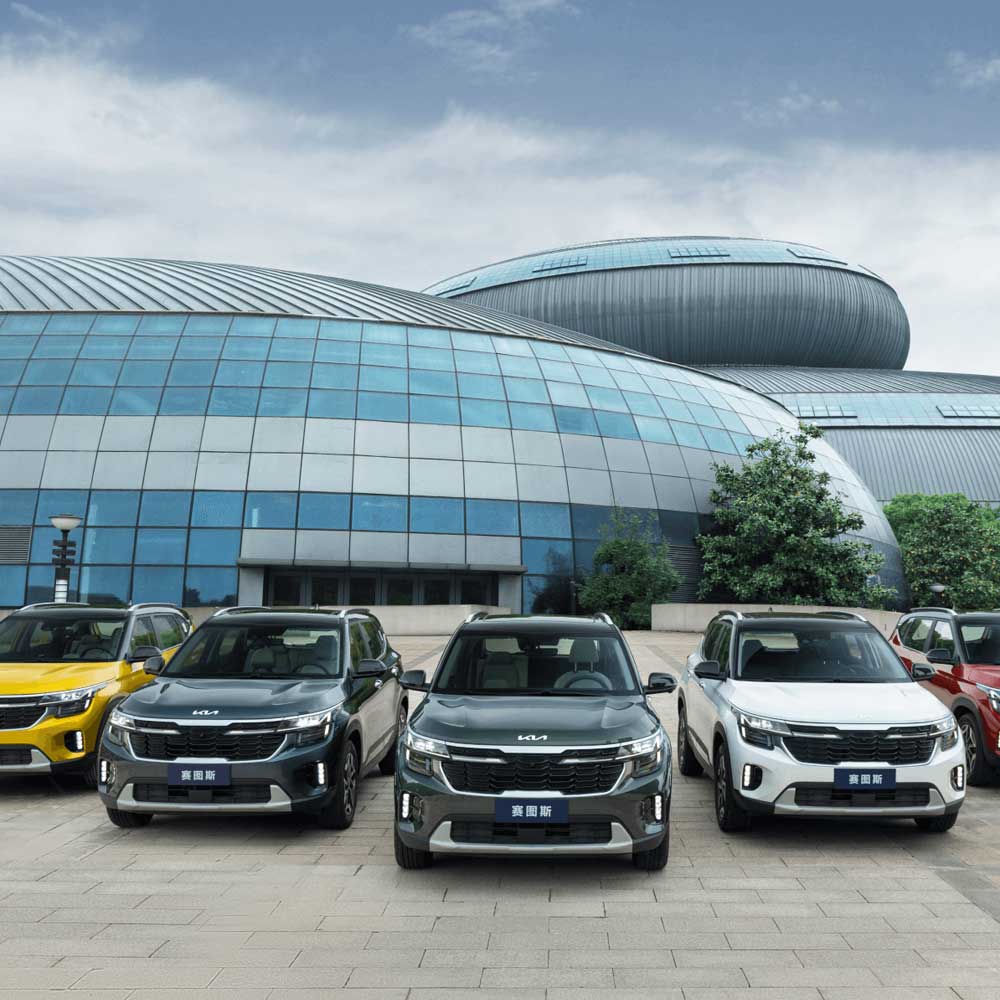 KIA Seltos 2023 1.5L CVT Premium Edition gallery2 Multiple Kia SUVs in different colors parked in front of a modern building with glass and metal architecture.