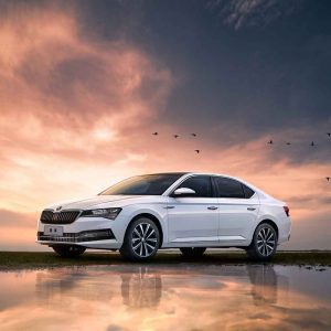 A white Škoda Superb sedan under a sunset sky with a reflection in water.