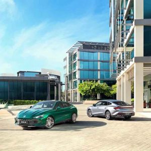 Two MG cars, one green and one silver, parked in a modern urban area with glass - walled buildings under a clear blue sky.