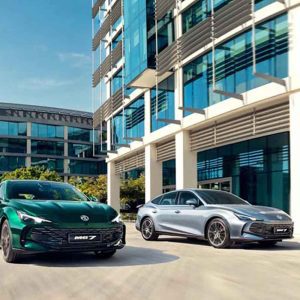 Two MG cars, one green and one silver, parked in front of a modern glass building under a clear blue sky.