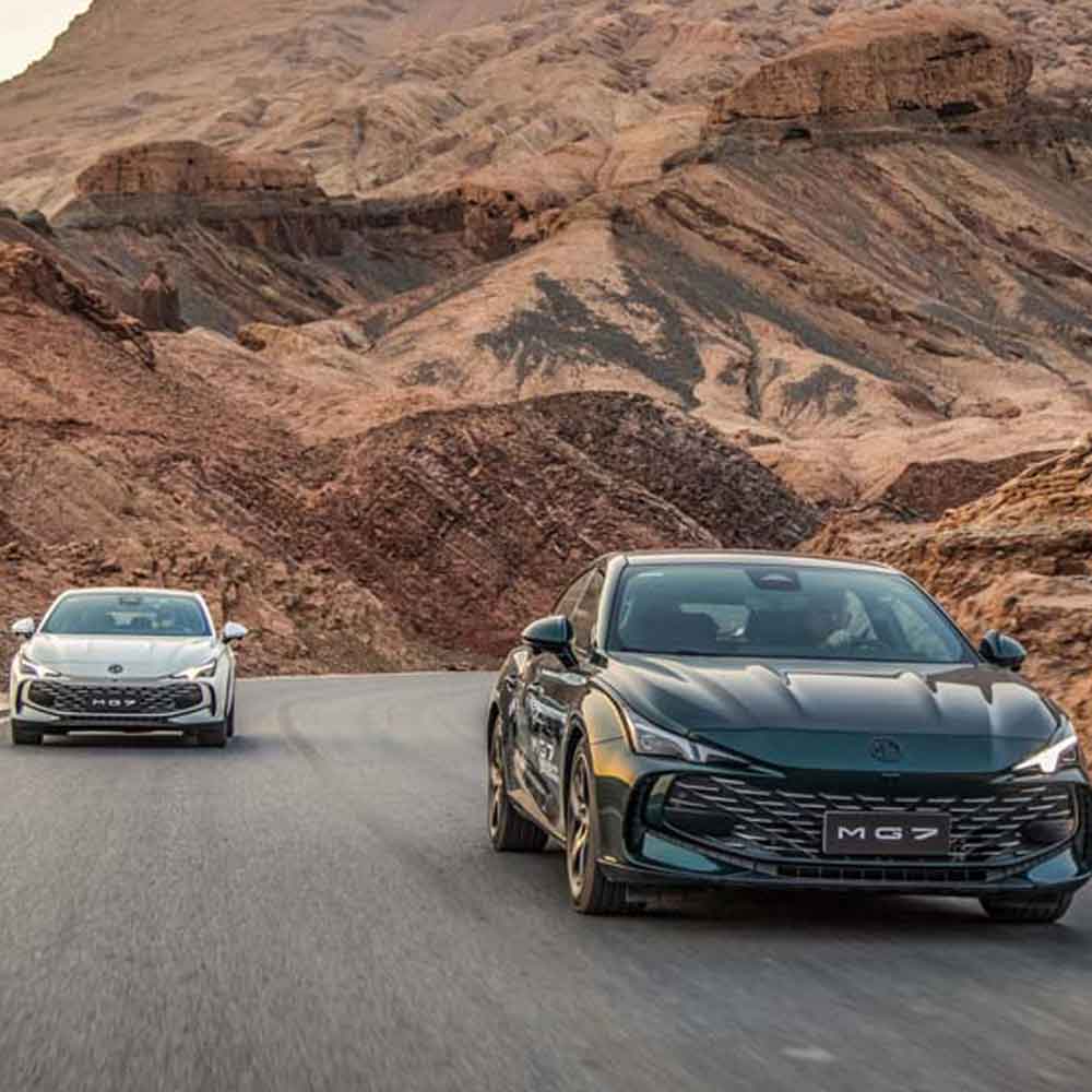 MG7 2025 1.5T 300DCT Luxury Edition product gallery image 1 Two MG7 cars, one dark green and one white, driving on a road with rugged, brown mountain scenery in the background.