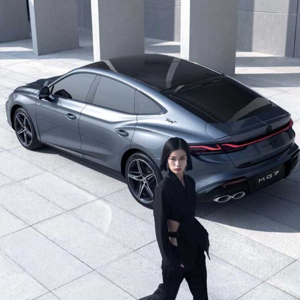 A woman in black clothing walks past a silver MG7 car in a modern, open - air architectural setting.