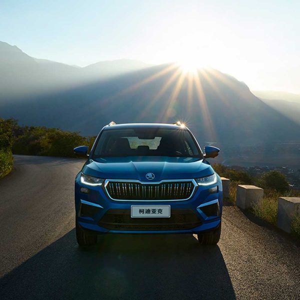 A blue Skoda Kodiaq SUV on a winding mountain road with the sun shining behind the mountains.