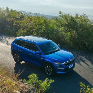 SKODA KODIAQ 2025 TSI330 5-Seater FWD Luxury Edition gallery image High Angle Shot A blue Skoda SUV driving on a winding road surrounded by lush vegetation with a scenic view in the background.