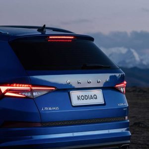 SKODA KODIAQ 2025 TSI330 5-Seater FWD Luxury Edition gallery image rear wing Rear view of a blue Skoda Kodiaq with its taillights on, set against a backdrop of mountains and a twilight sky.