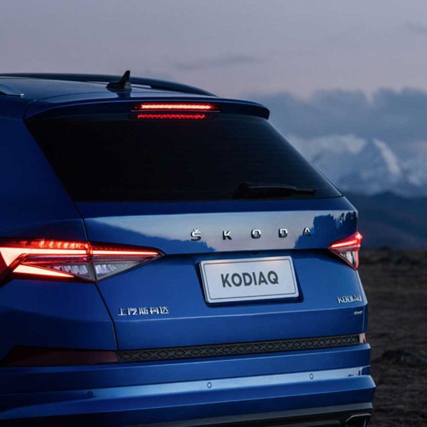 Rear view of a blue Skoda Kodiaq with its taillights on, set against a backdrop of mountains and a twilight sky.