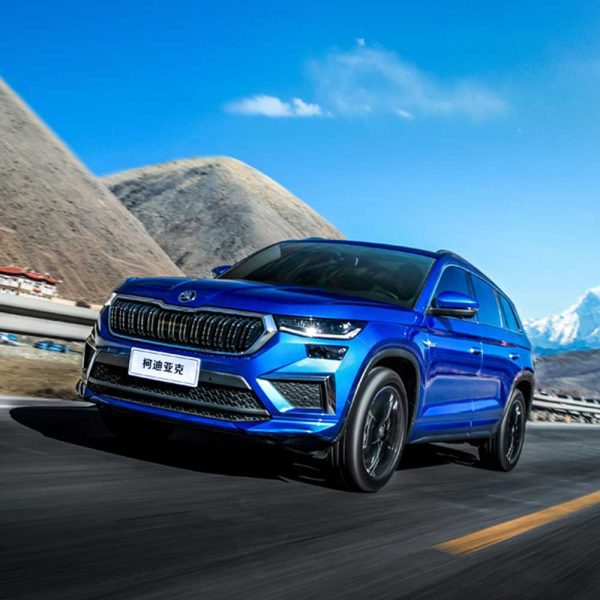 A blue Skoda Kodiaq SUV driving on a road with mountains and a clear blue sky in the background.