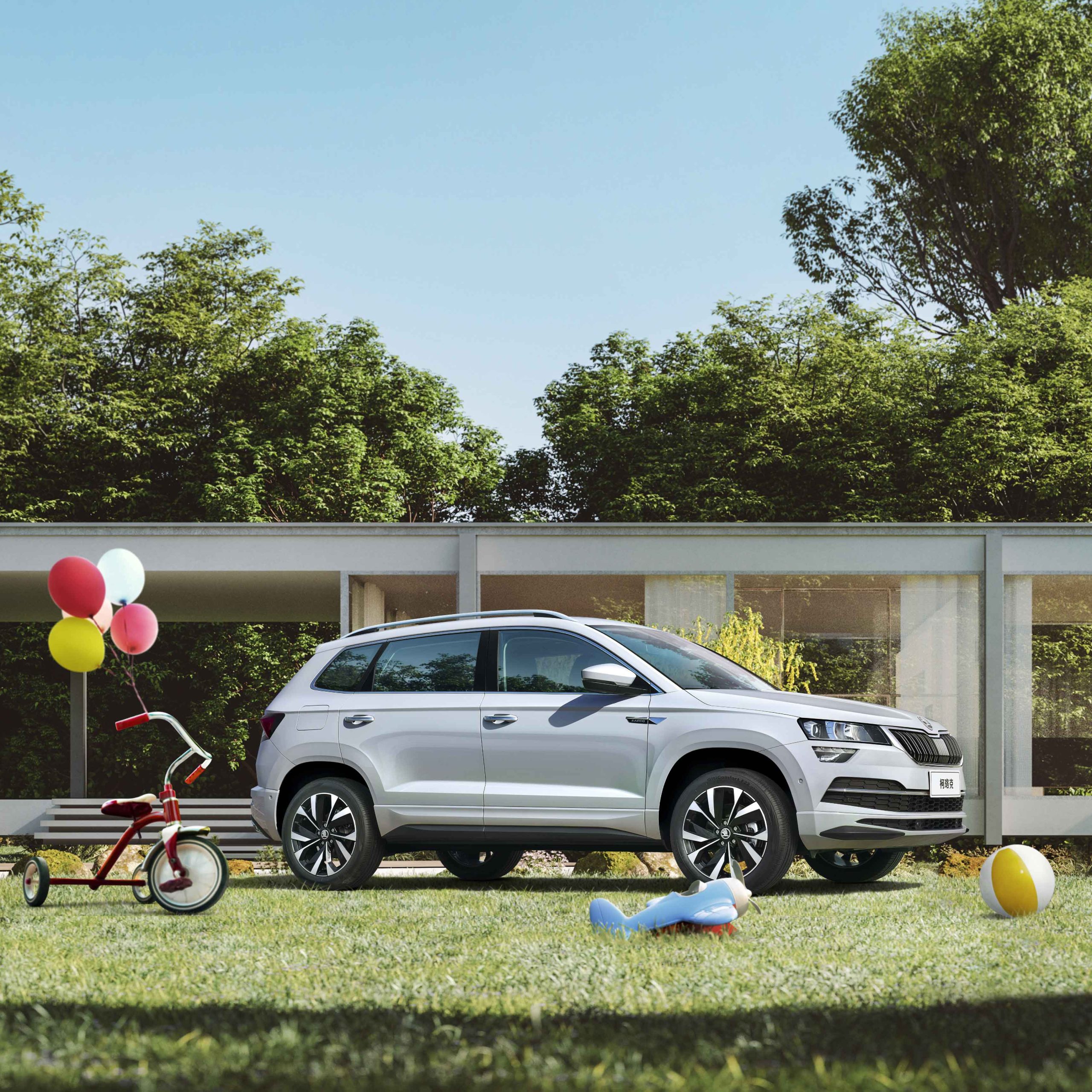 A silver Skoda SUV parked on a lawn, with children's toys like a tricycle and balloons around, in front of a modern house.