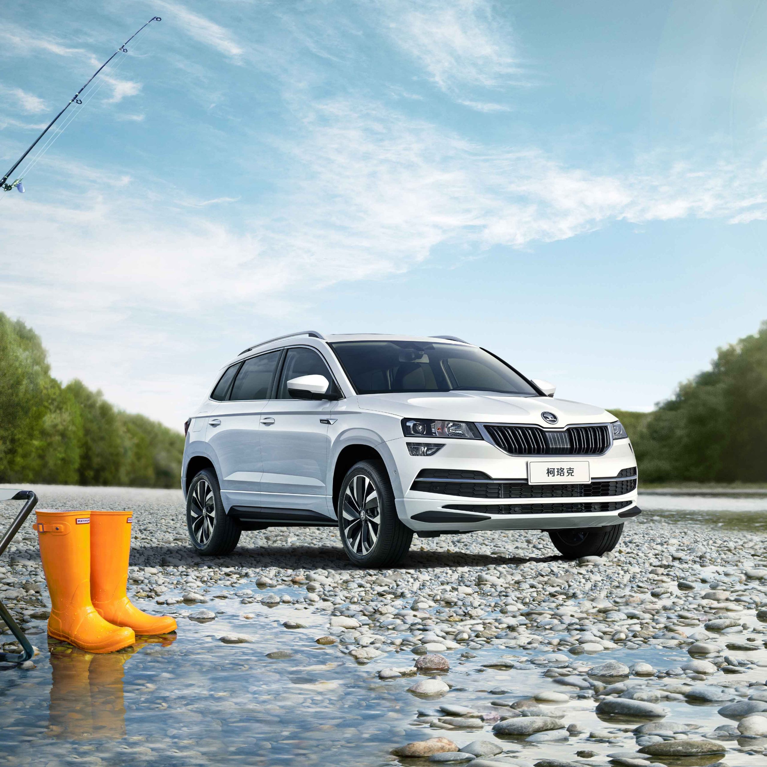 A white Skoda Karoq SUV parked on a rocky riverside, with orange rain boots and a fishing rod nearby.