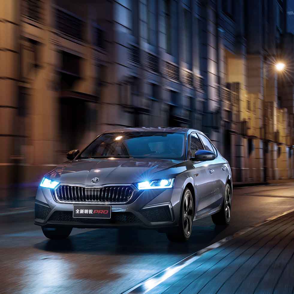 A silver Skoda Octavia Pro car driving on a city street at night, with buildings and lights in the background.