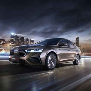 A brown Skoda car driving on a road with a cityscape in the background at dusk.