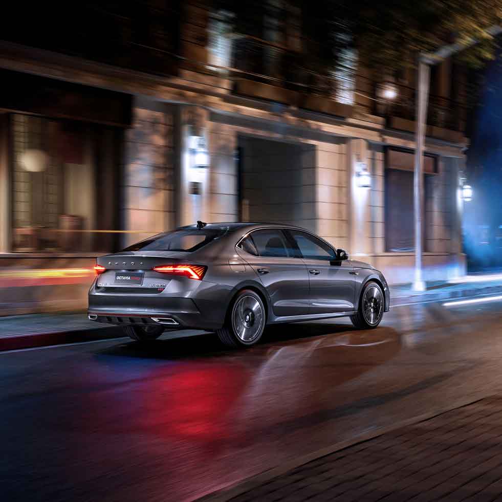 A silver Skoda Octavia sedan driving on a city street at night, with buildings and lights in the background.