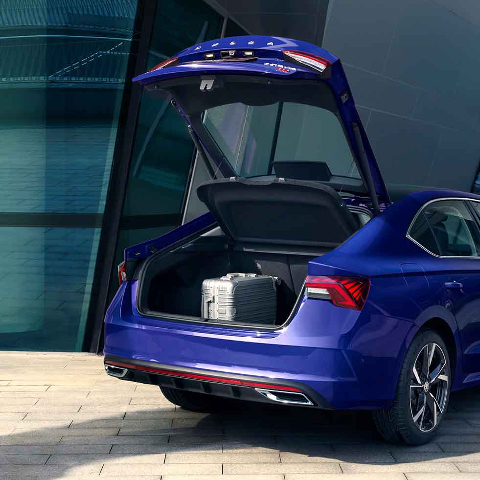 Rear view of a blue sedan with its trunk open, containing a silver suitcase, parked in front of a modern building.