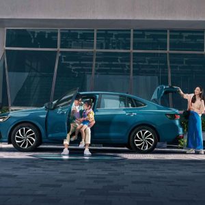 A family with a teal sedan, where a man holds a child by the open door and a woman stands by the open trunk.
