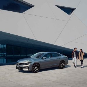 A silver Volkswagen sedan parked in front of a modern building, with two people walking nearby.