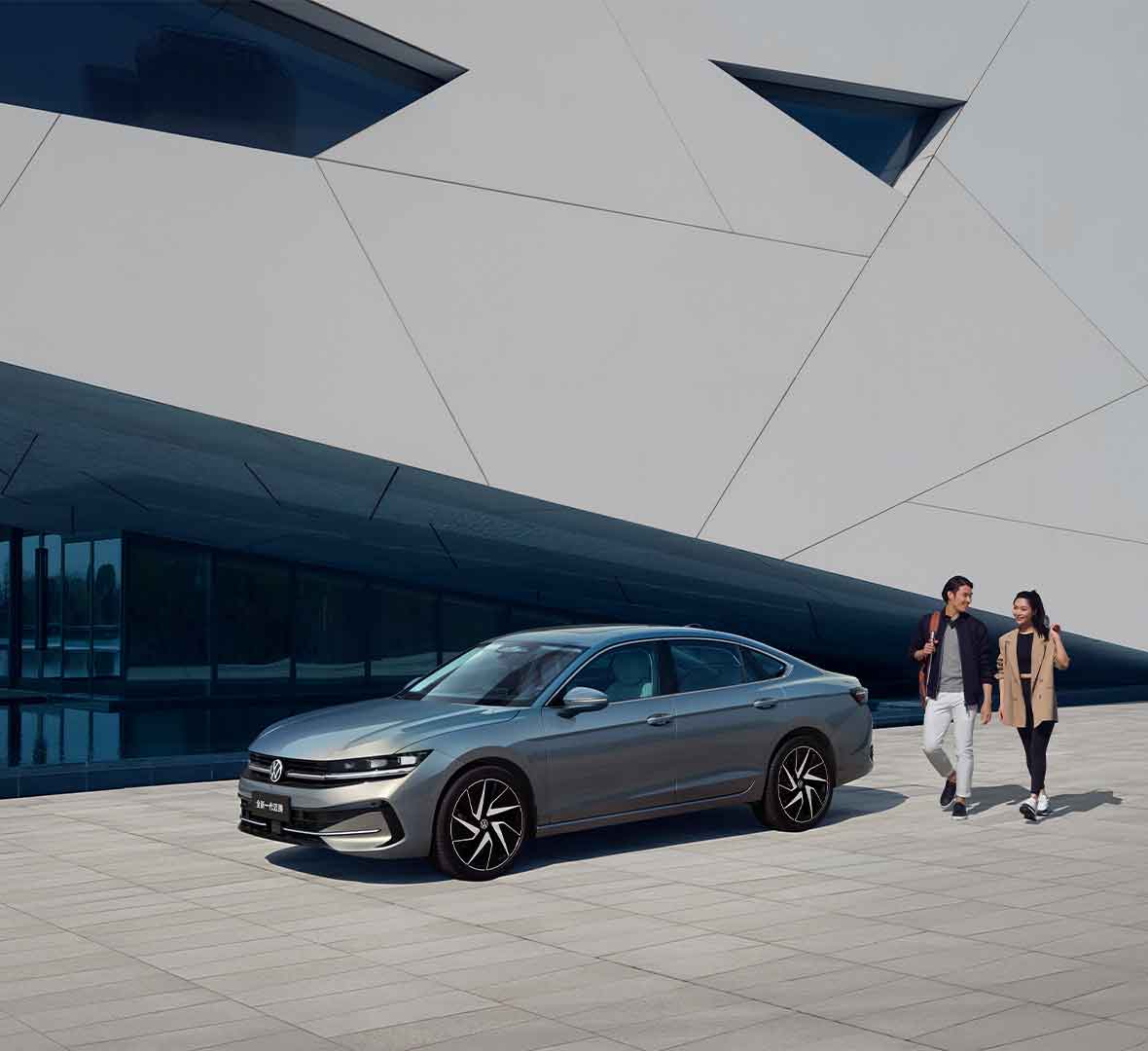 A silver Volkswagen sedan parked in front of a modern building, with two people walking nearby.