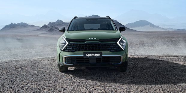 Front view of a green Kia Sportage SUV parked in a desert-like, mountainous landscape. Features the Kia logo, bold grille, and "Sportage" badging on the front, set against a vast, arid backdrop with distant peaks.