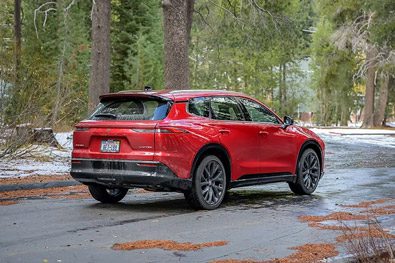 Rear view of a vibrant red SUV parked on a wet, pine-strewn forest road with patches of snow.