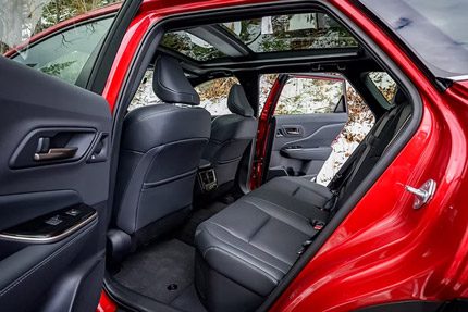 Rear passenger view of a red SUV, featuring black leather seats and a panoramic sunroof with trees visible outside.
