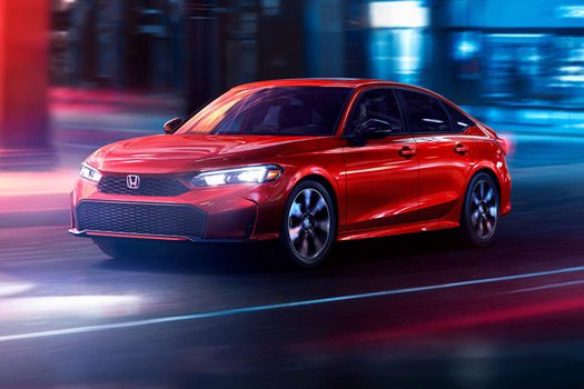 2026 Honda Civic Sedan Sport Touring Hybrid in Rallye Red front three-quarter view driving through a city